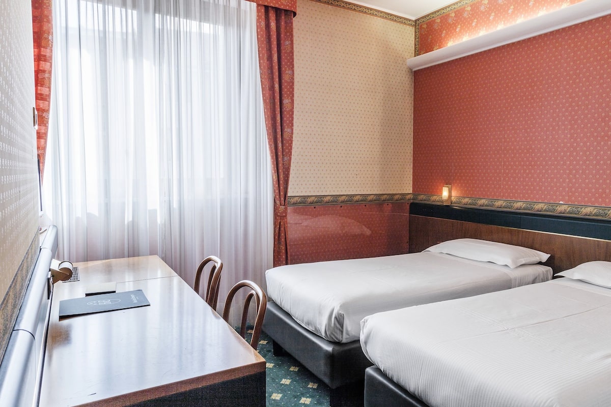 A hotel room with two beds, a wooden desk, a chair, and a window with curtains.