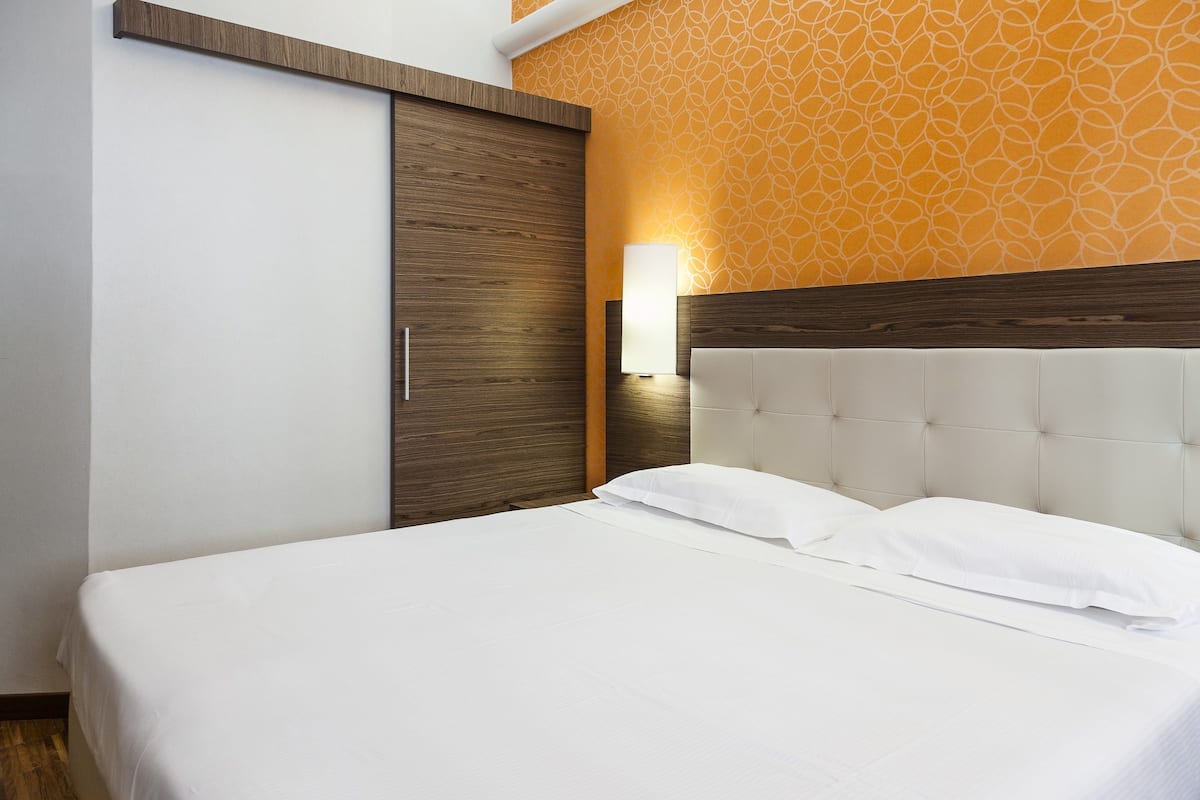 A neatly made bed with white linens in a room with a wooden wardrobe and a patterned wall.