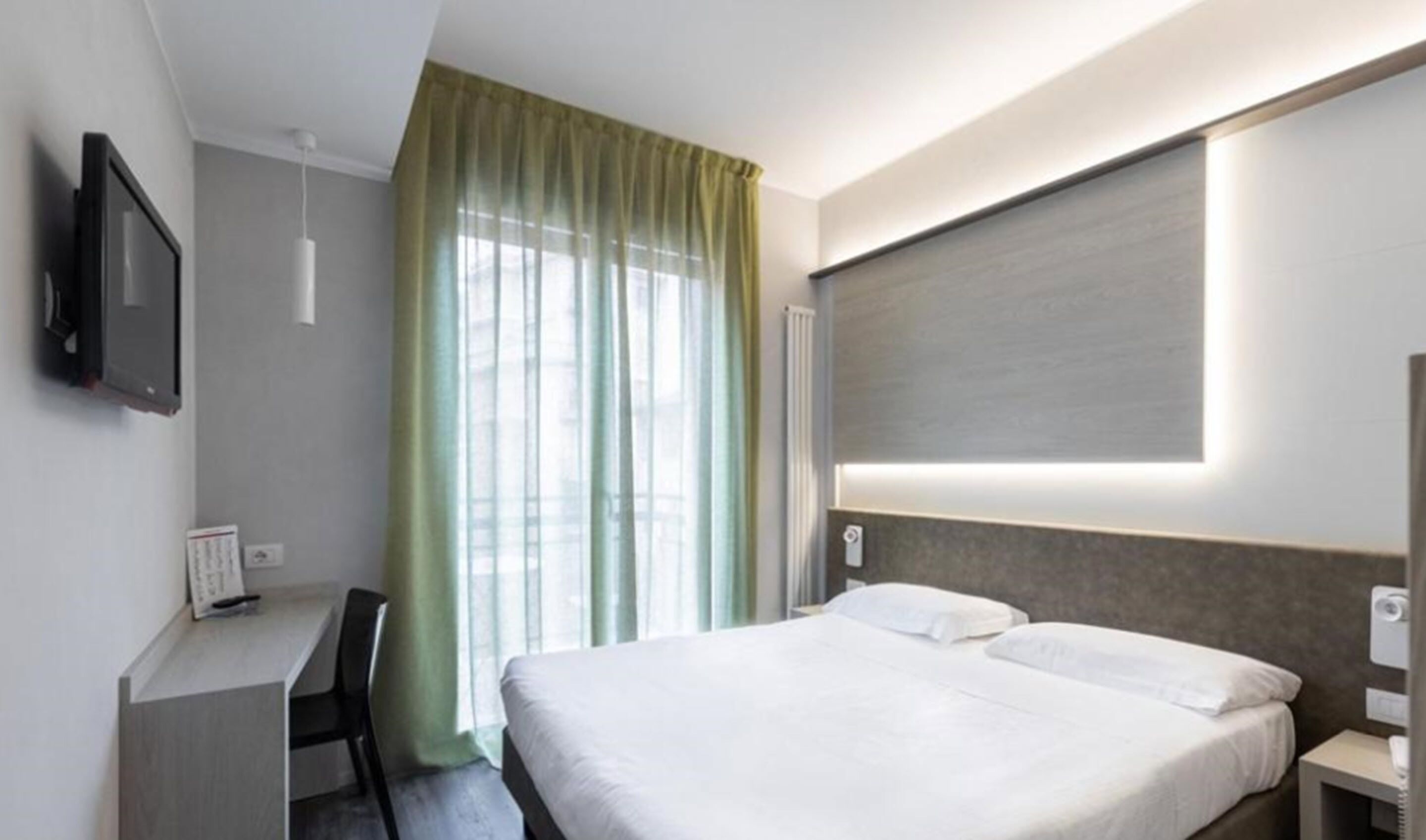 Habitación de hotel con cama, escritorio, silla, televisor y ventana con cortinas.