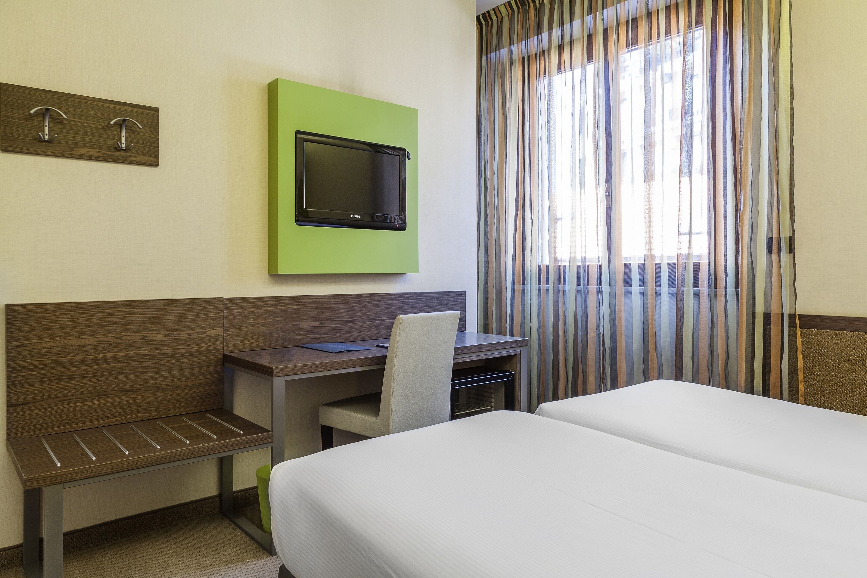 Een hotelkamer met een bed, een bureau met stoel, een televisie bevestigd aan een groen paneel, en een raam met gestreepte gordijnen.