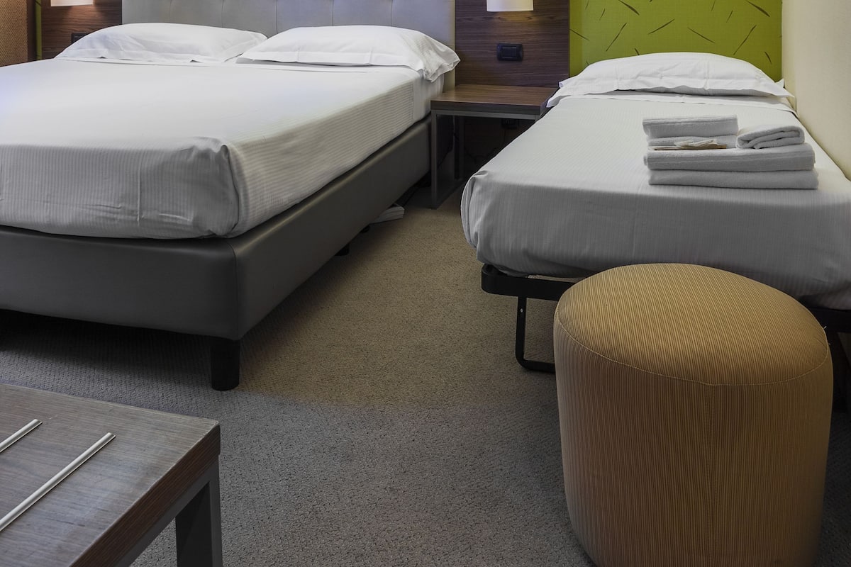 A hotel room with two beds, a nightstand, a lamp, and a small ottoman.