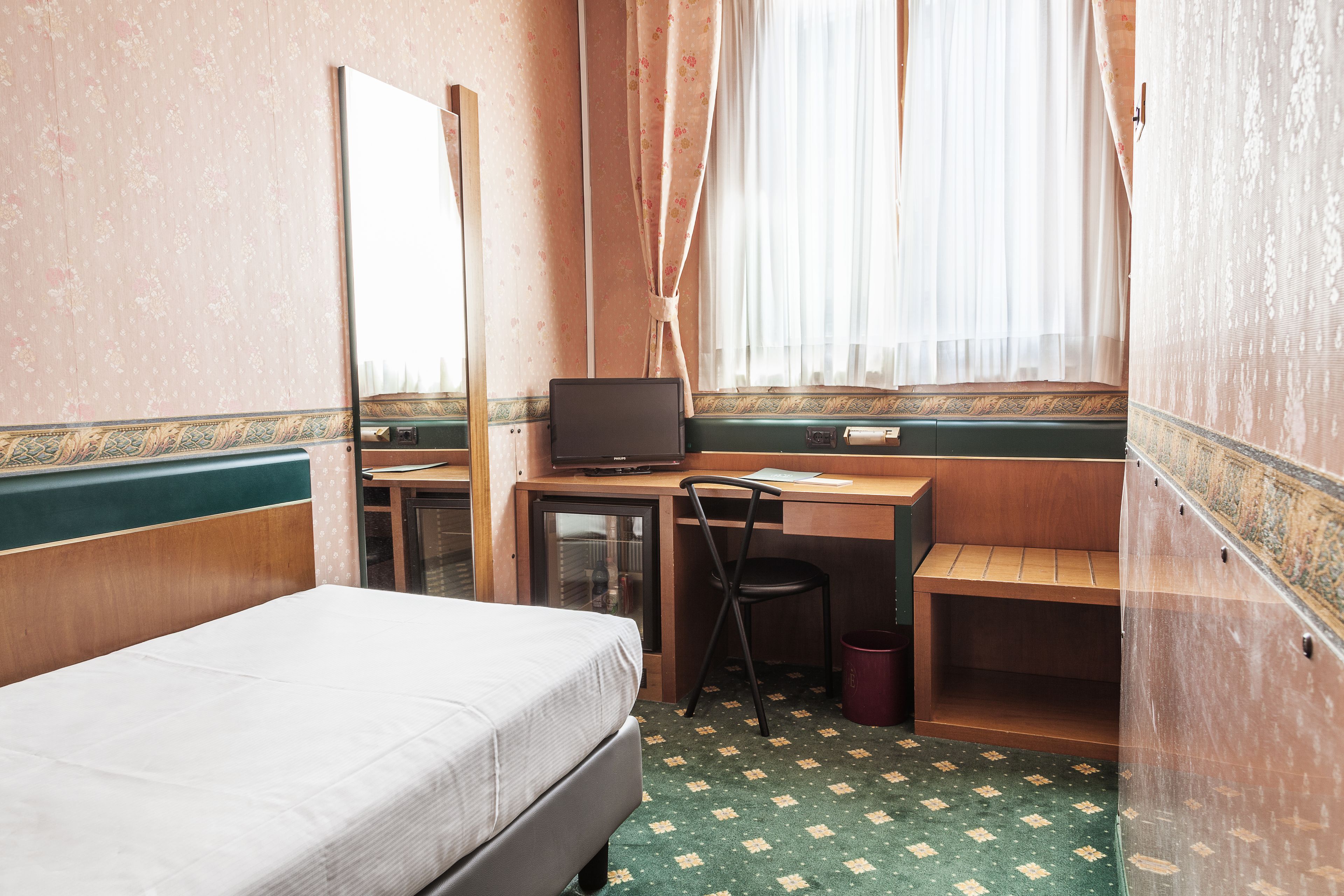 Ein kleines Hotelzimmer mit einem Einzelbett, einem Schreibtisch mit Computer, einem Stuhl und einem kleinen Fenster mit transparenten Vorhängen.