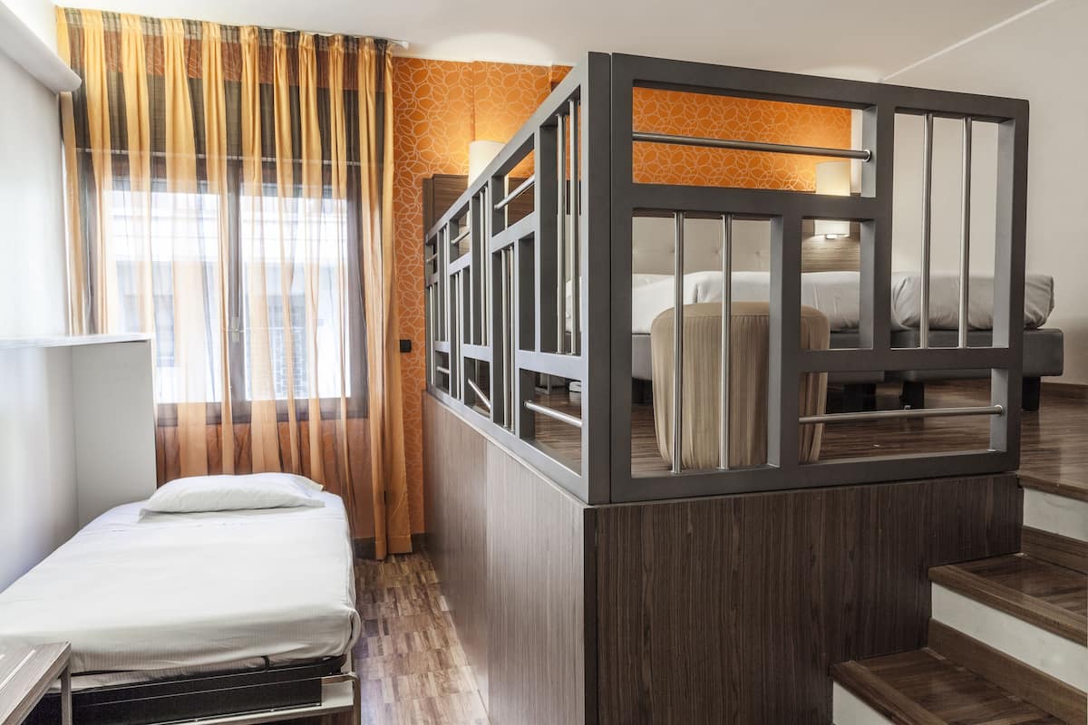 A hotel room with a bed, a desk, a window with curtains, and a staircase.