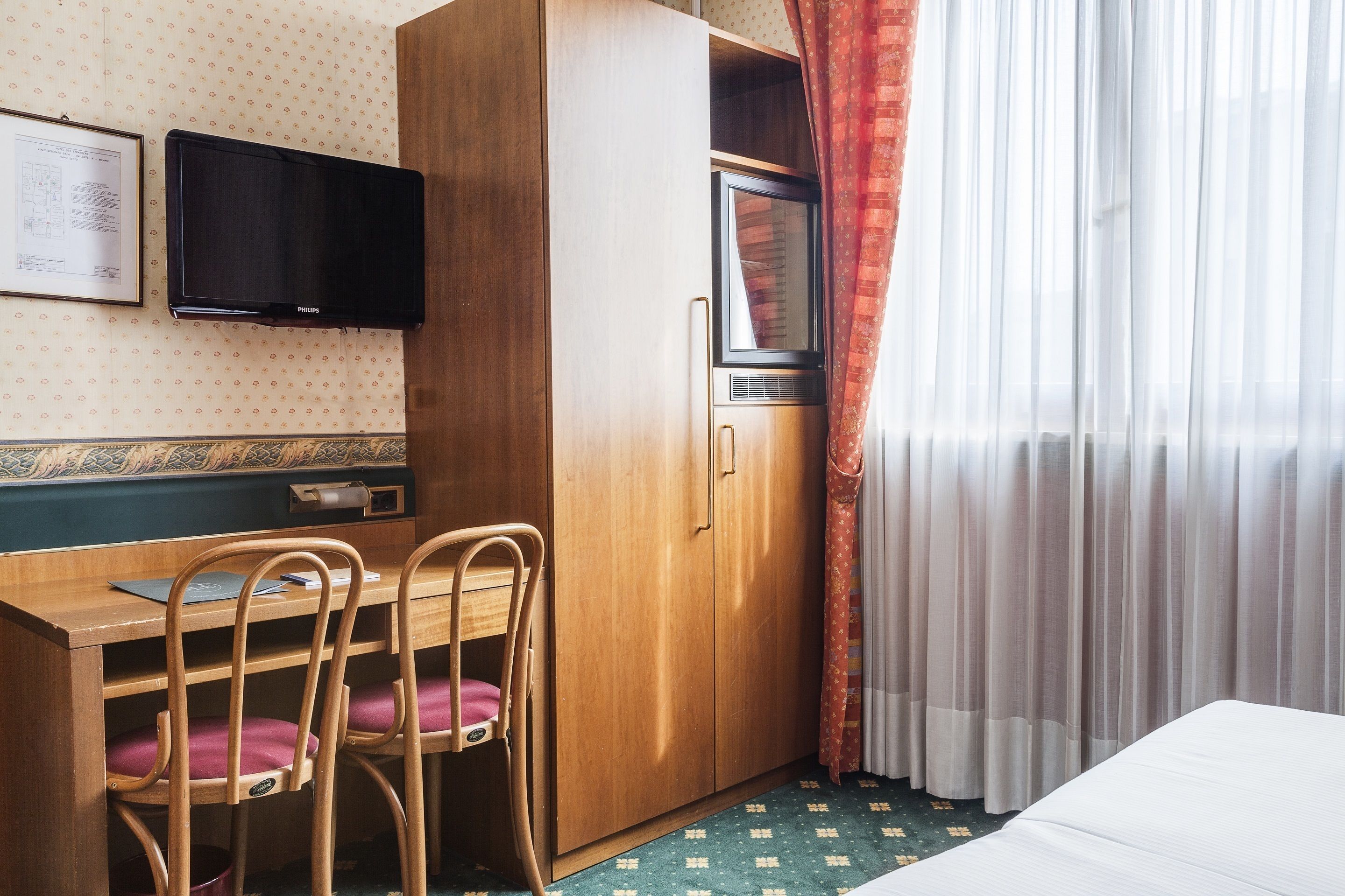 A hotel room with a wooden desk, two chairs, a television, a wardrobe, and a window with curtains.
