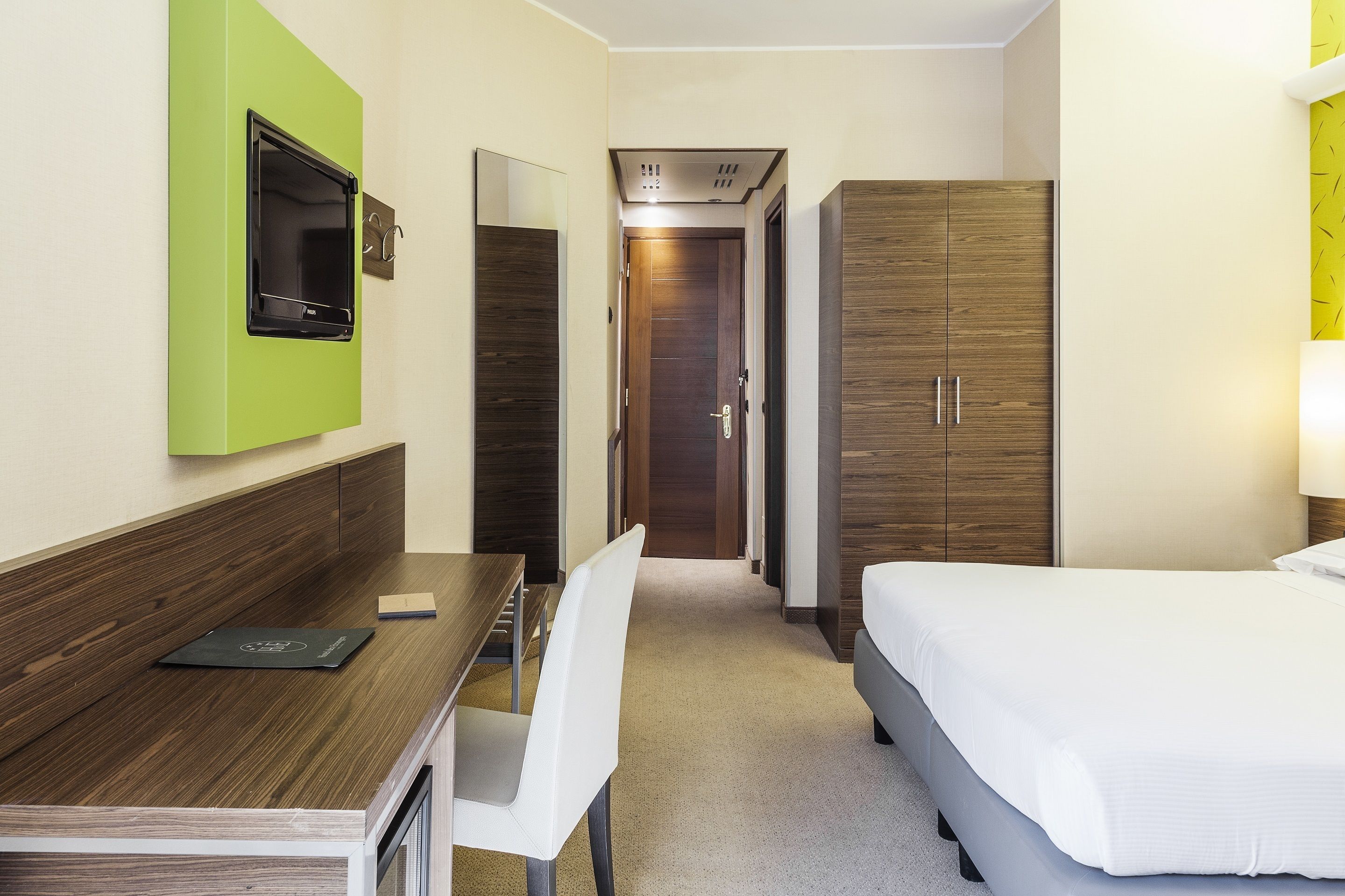 Une chambre d’hôtel comprenant un lit, un bureau avec une chaise, un téléviseur fixé sur un mur vert et une armoire.