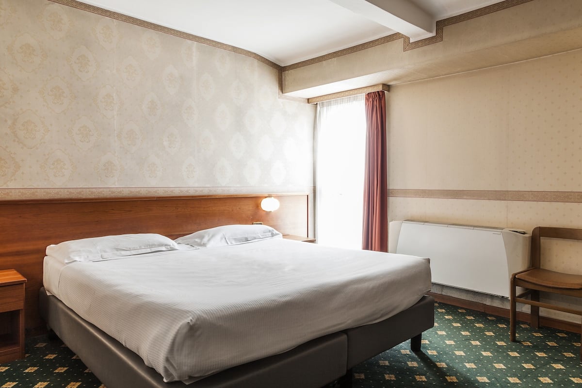 A hotel room with a large bed, a wooden headboard, a nightstand, a chair, a window with curtains, and a radiator.