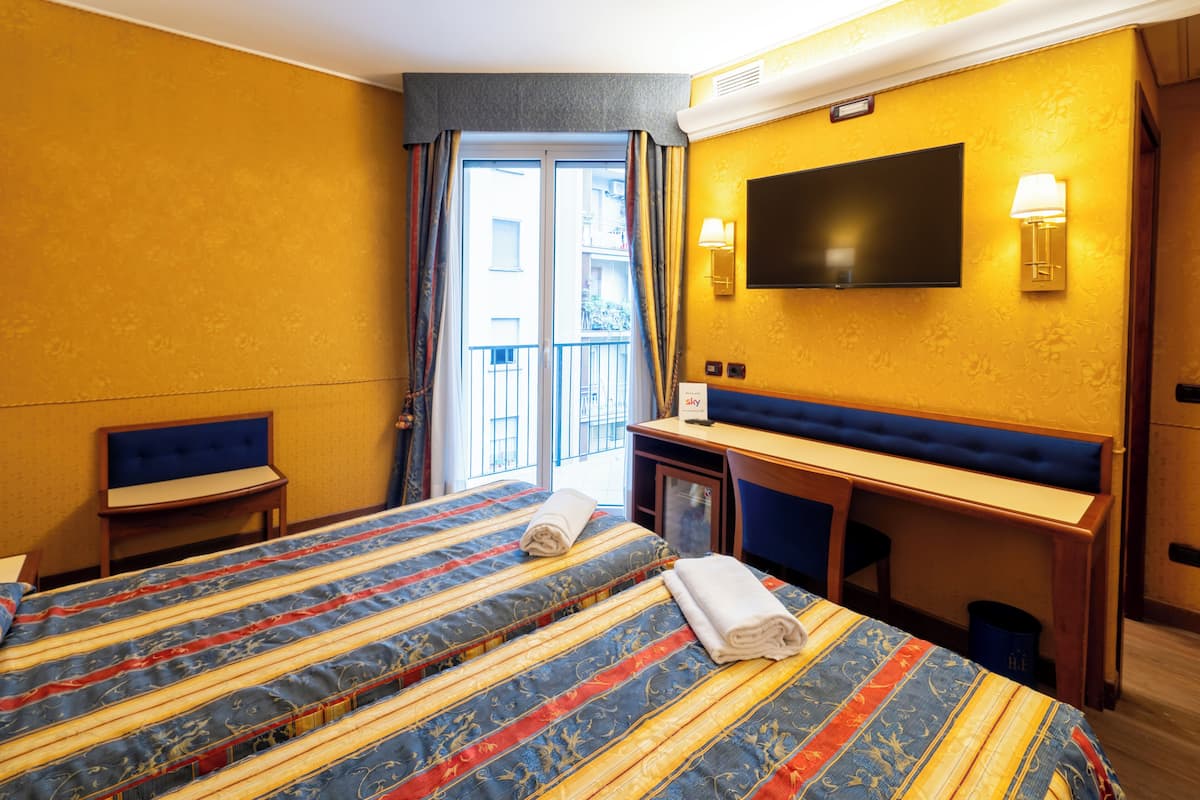 A hotel room with a large bed, a desk with a chair, a TV mounted on the wall, and a window with curtains.