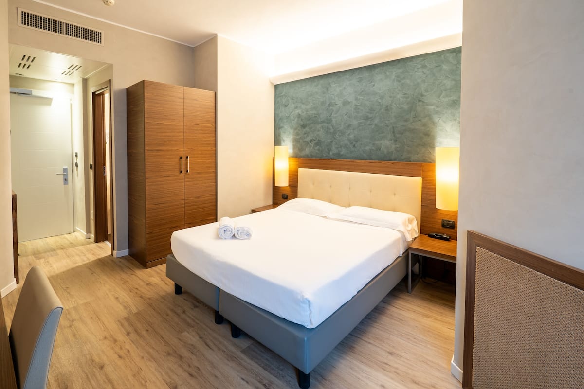 A hotel room with a bed, wooden wardrobe, bedside lamps, and a headboard with a textured wall above.