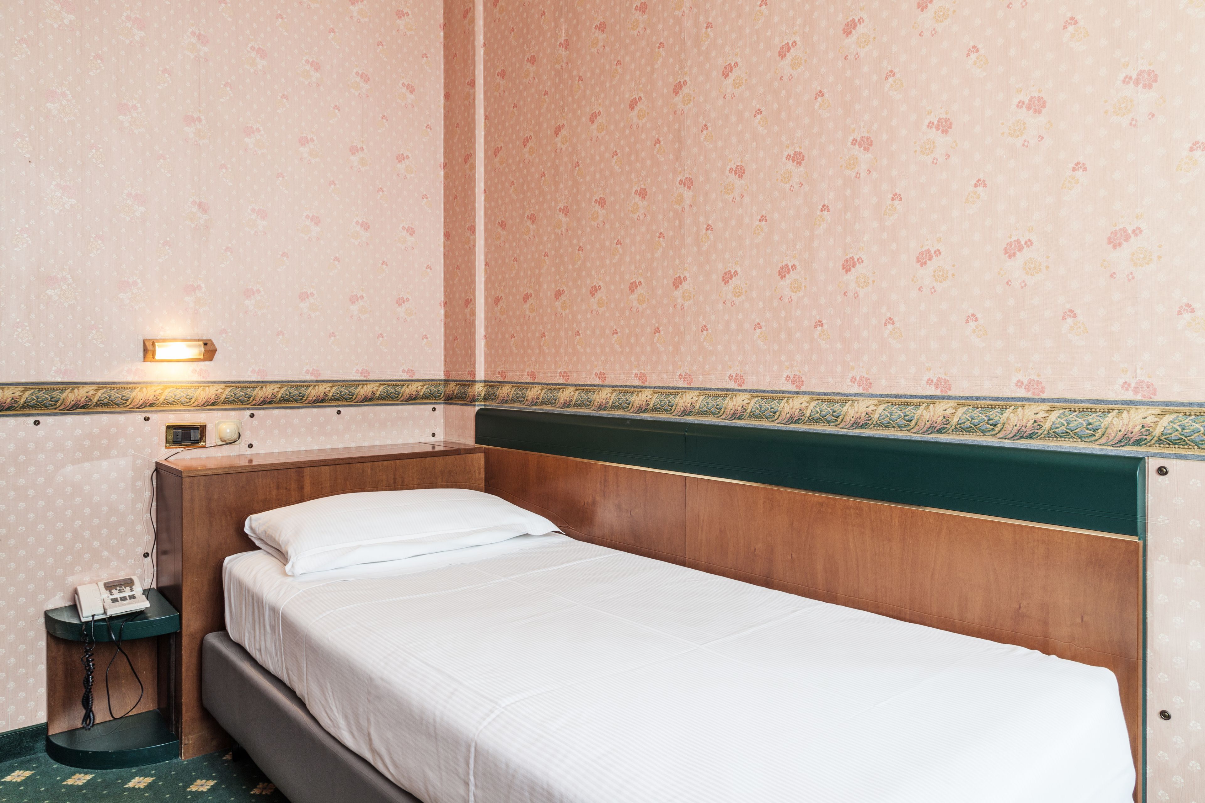 Una cama individual con cabecera de madera, una mesita de noche con lámpara y un teléfono.
