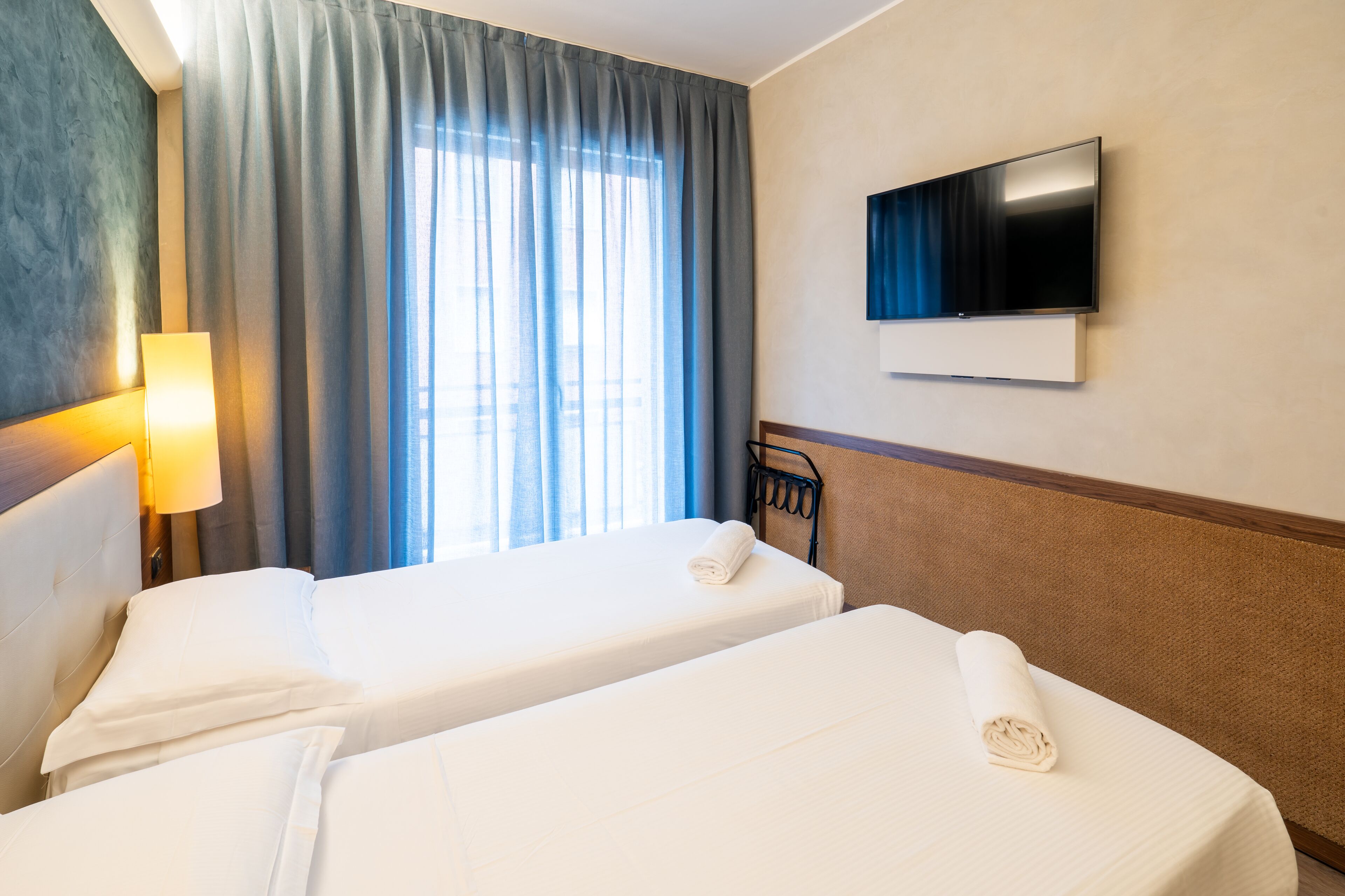 A hotel room with two beds, a TV on the wall, and a window with curtains.