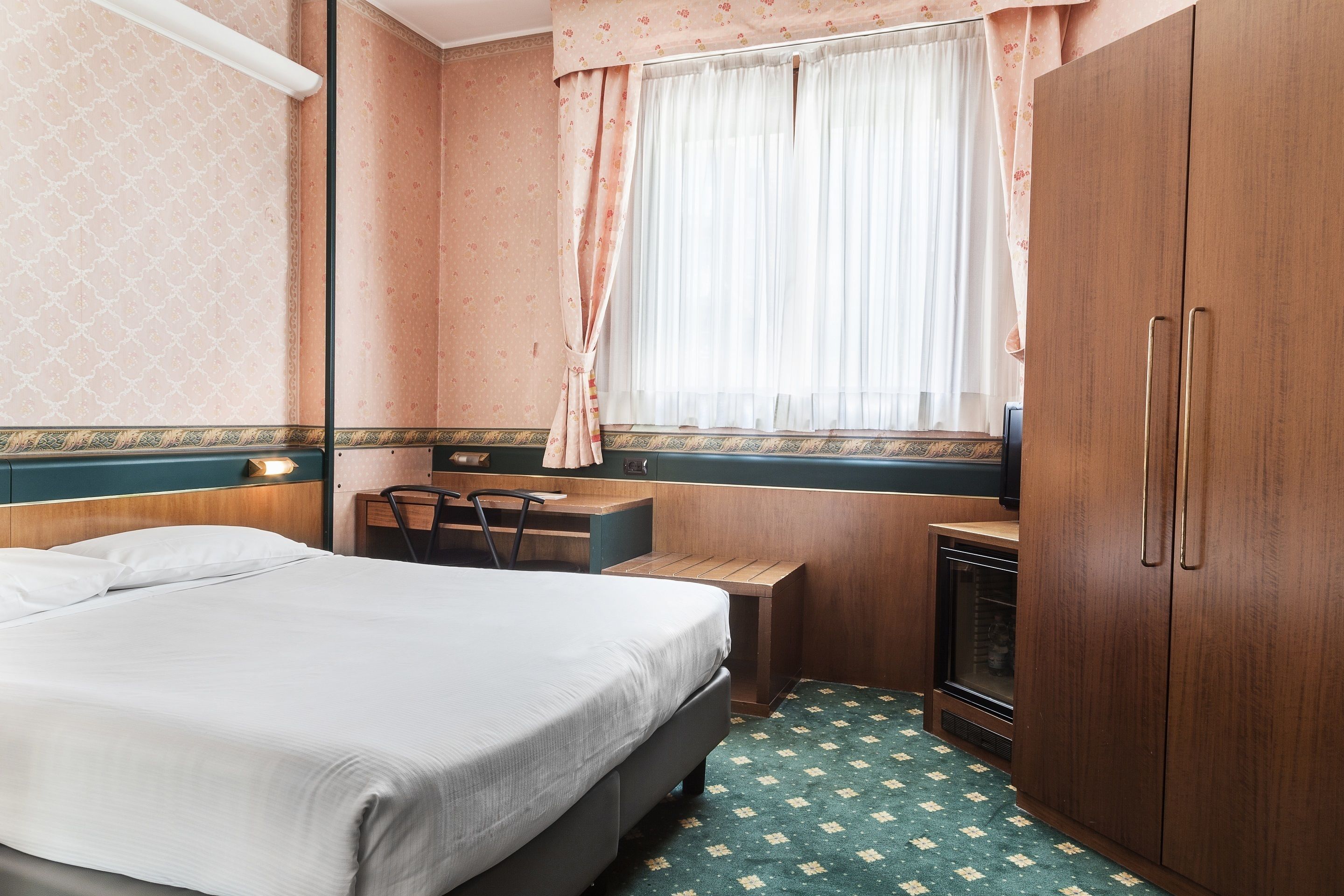 Une chambre d’hôtel de taille modeste, comprenant un lit, un bureau, une chaise, une armoire et une fenêtre avec des rideaux.