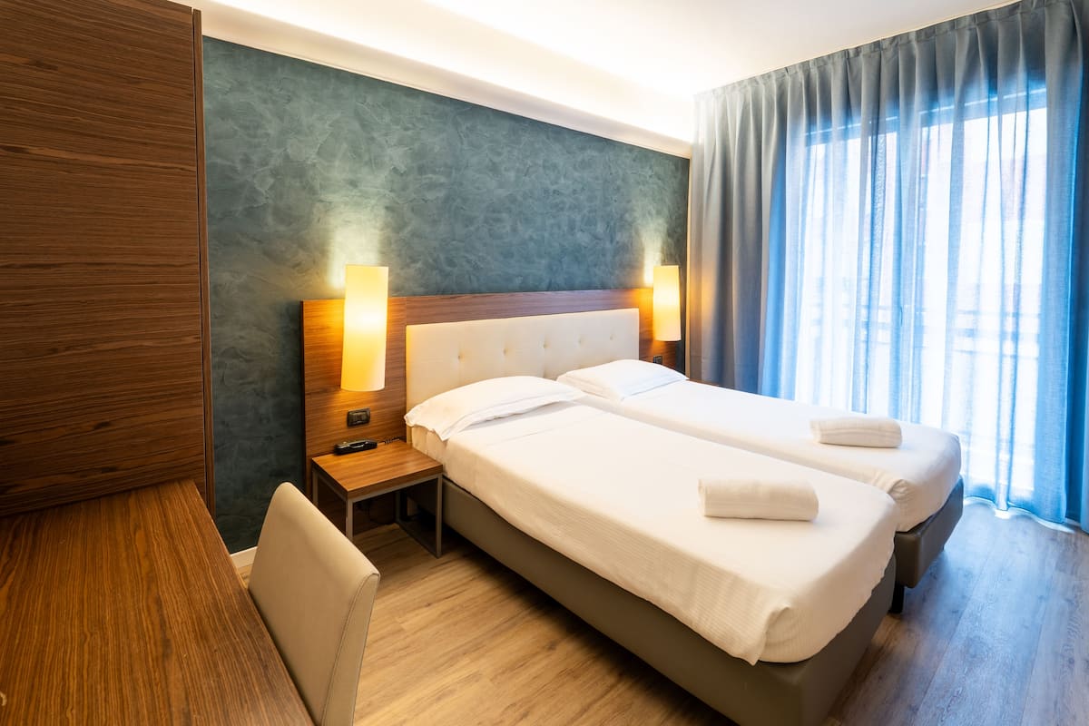 A hotel room with two beds, wooden headboard, bedside tables with lamps, and a view of the outdoors through large windows.