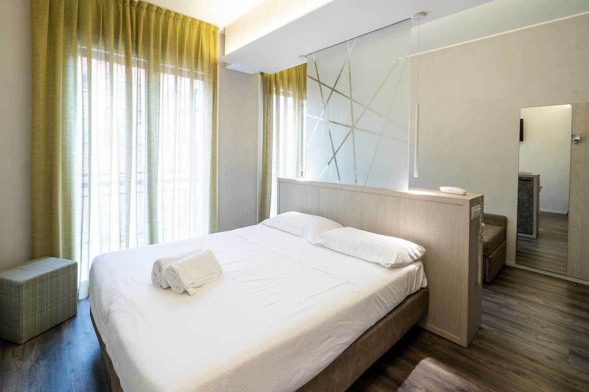 A modern hotel room with a large bed, a bedside table, a mirror, and a small seating area.