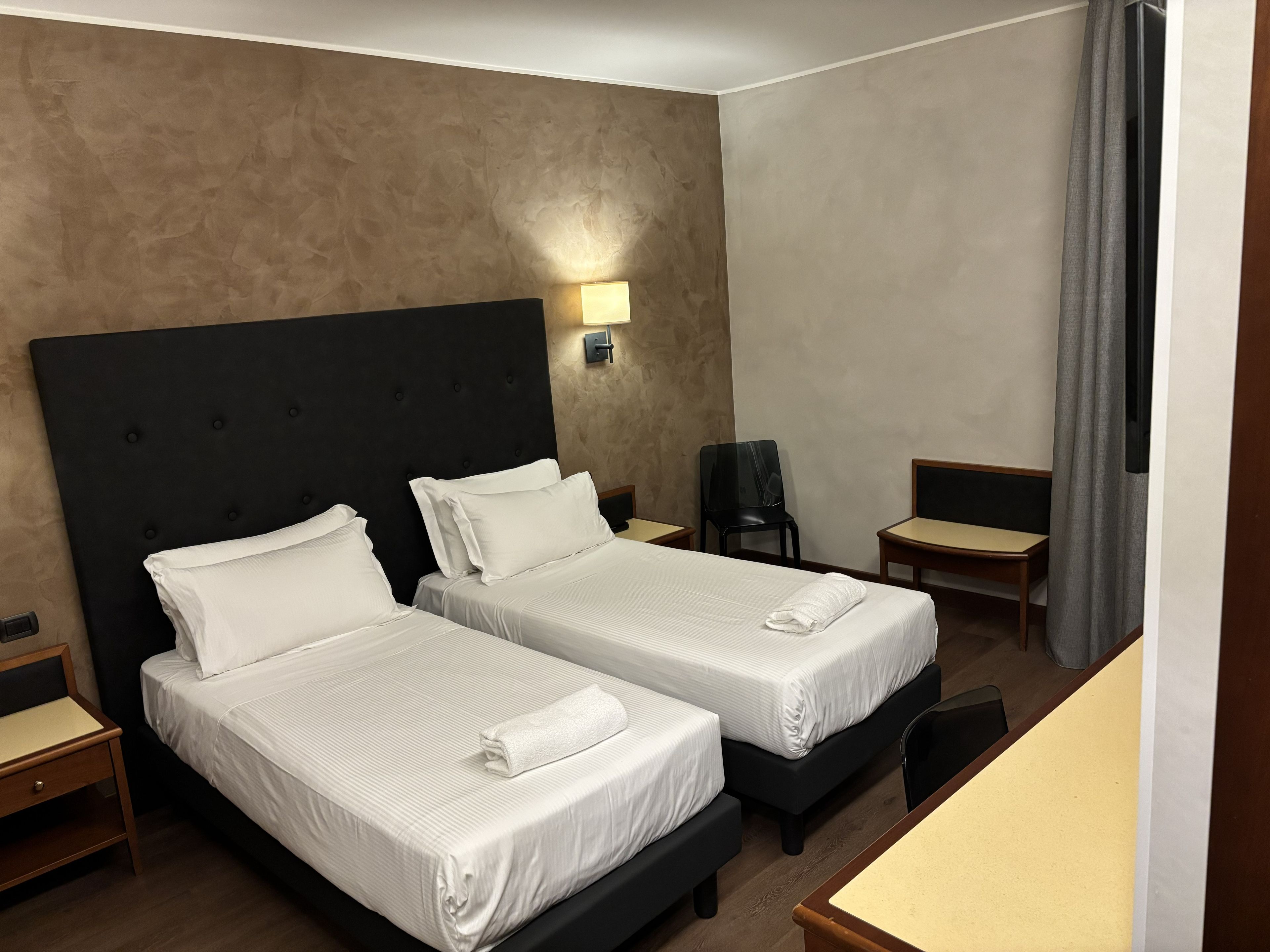 A hotel room with two beds, a desk, a chair, and a wall-mounted lamp.