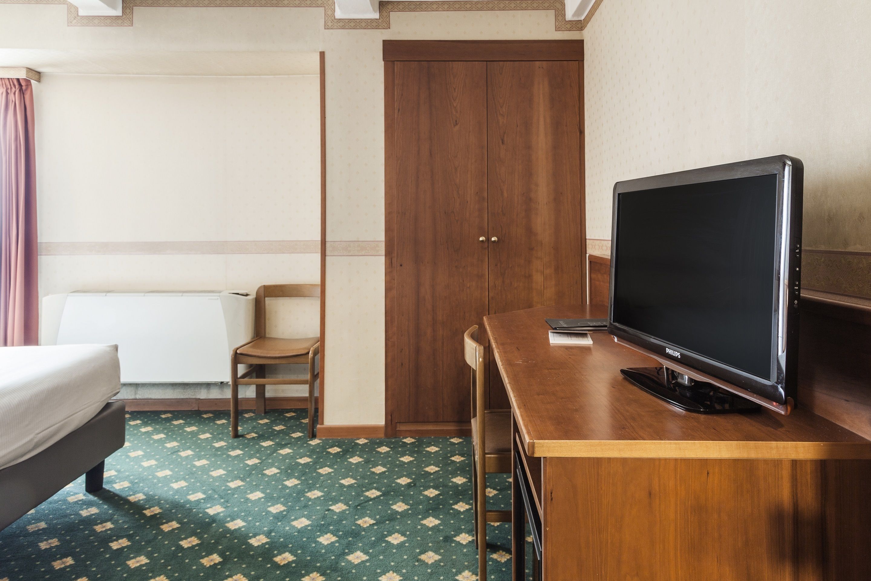 Une chambre d’hôtel comprenant un lit, un bureau en bois avec une télévision, une chaise et une armoire.