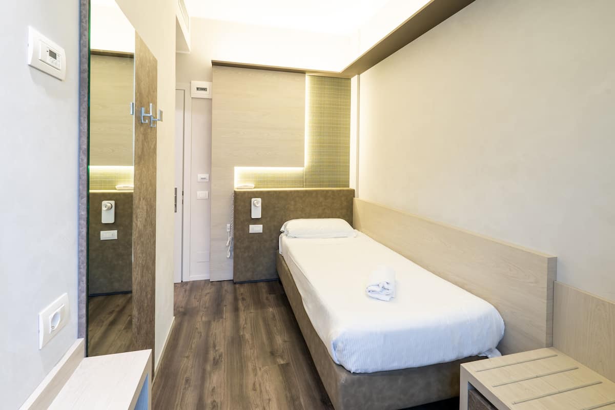 A small, modern hotel room with a single bed, a bedside table, a mirror, and a small window.