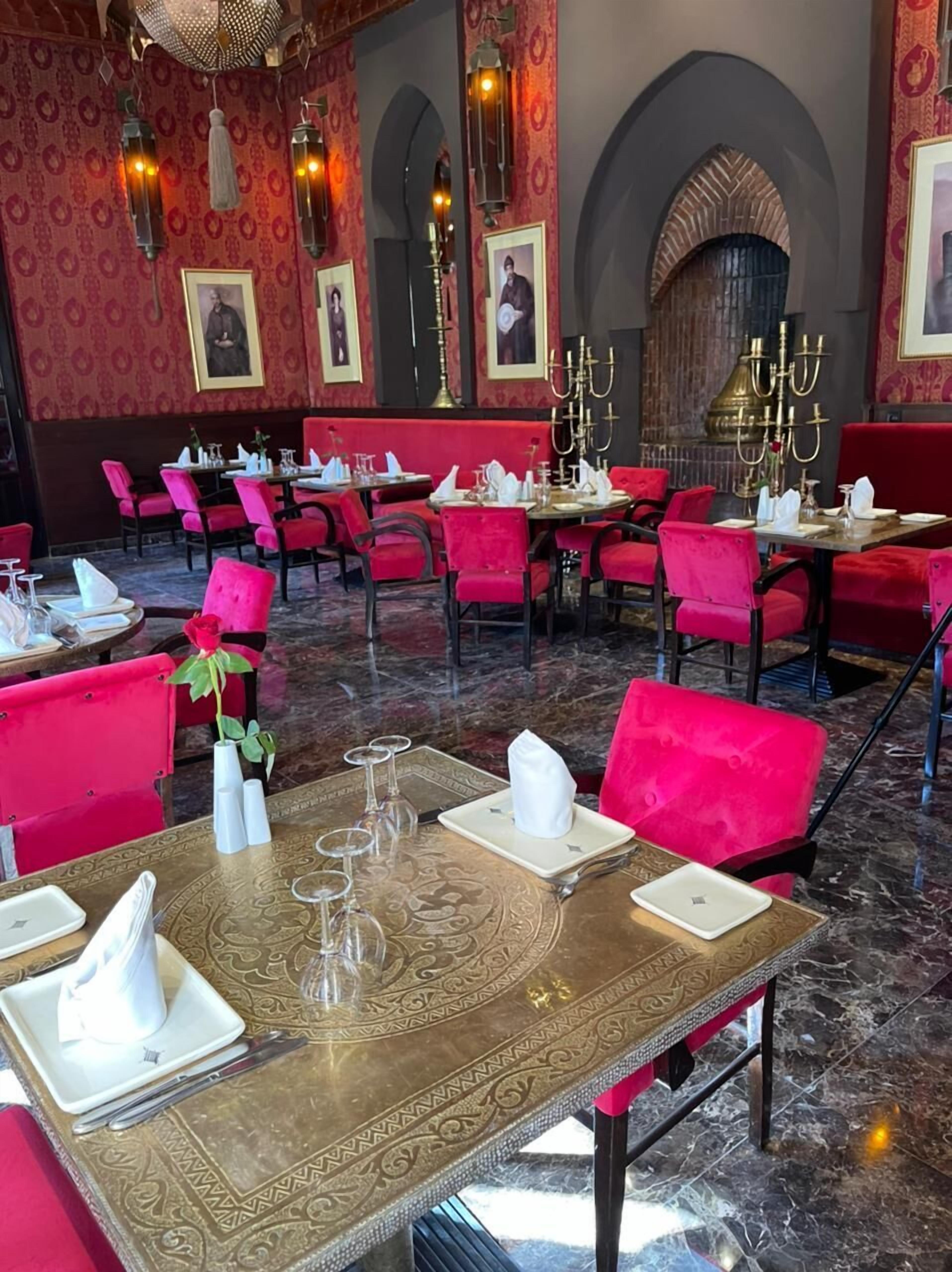 Um restaurante com cadeiras estofadas em vermelho, mesas elaboradas e papel de parede estampado.