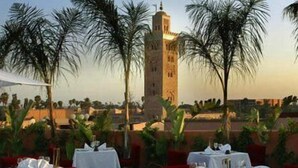 Un área de comedor al aire libre con mesas dispuestas para una comida, palmeras y una torre al fondo.