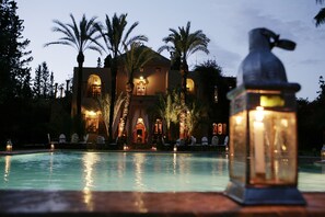 Uma área de piscina com um edifício grande ao fundo, palmeiras e um candeeiro decorativo.