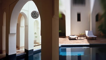 Zona de piscina con una silla de descanso y una mesa, con vista a un edificio de arcos y una decoración esférica colgante.