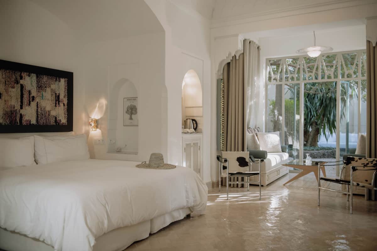 A spacious bedroom with a large bed, a seating area with a sofa and chairs, and a view of greenery through a window.