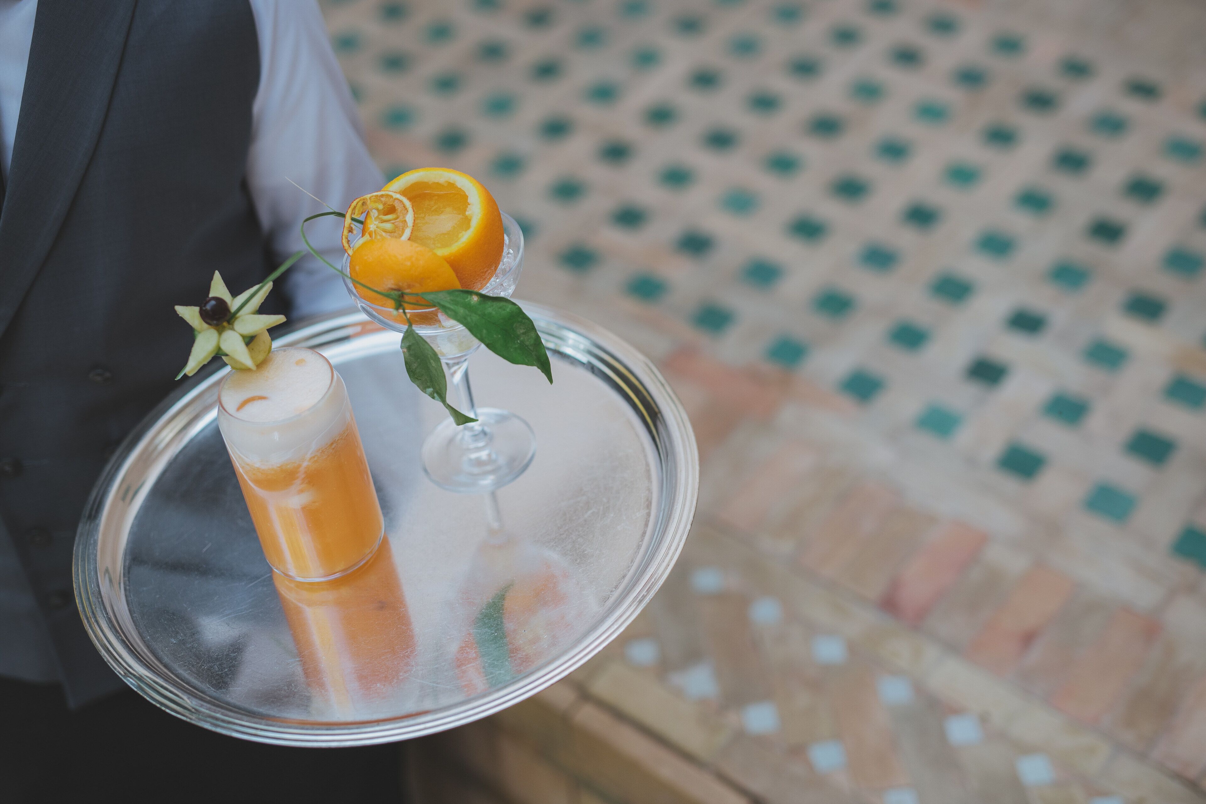 A tray with a glass of orange juice and a cocktail, garnished with an orange slice and a leaf.