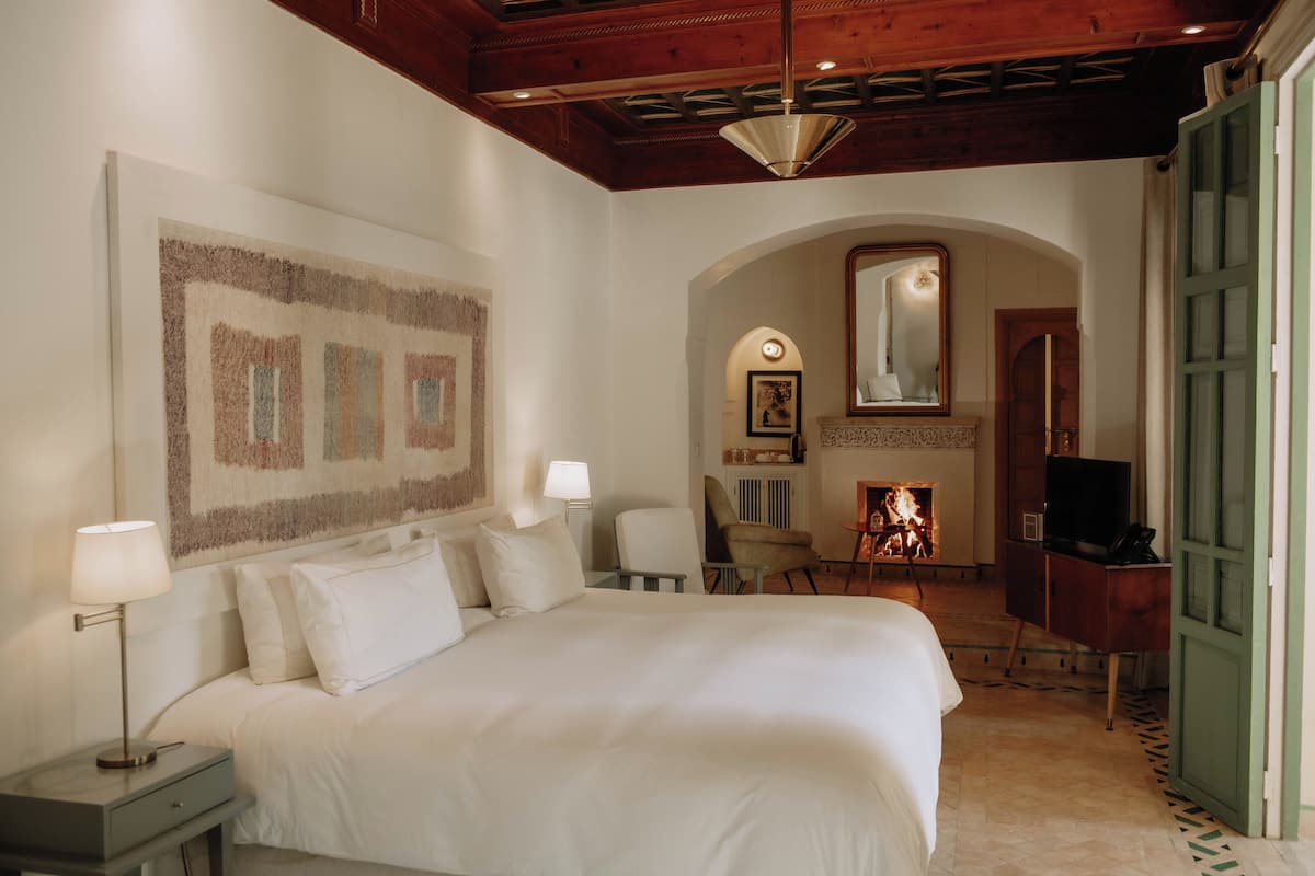 A hotel room with a large bed, a desk with a lamp, a chair, a television, and a fireplace.