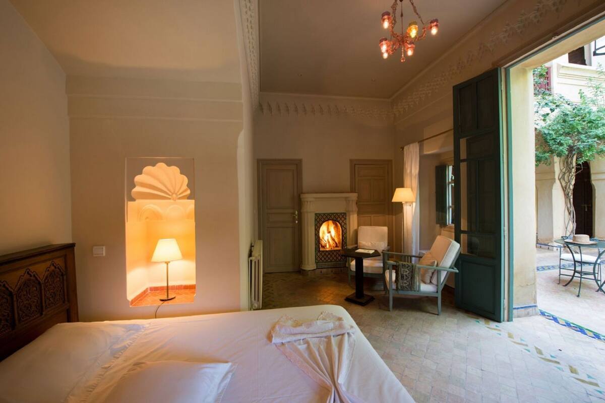 A hotel room with a bed, a chair, a table, a fireplace, and a view of the outside.