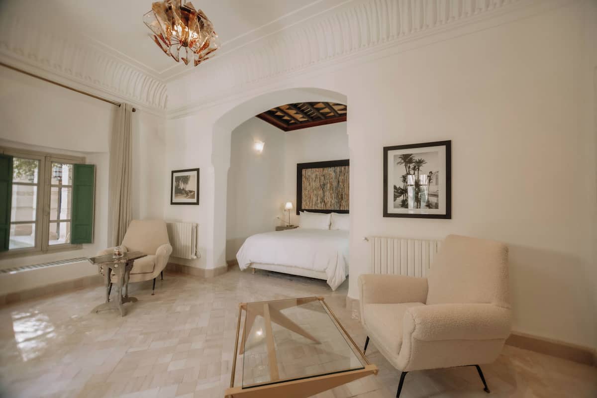A hotel room with a bed, two armchairs, a glass coffee table, a chandelier, and framed artwork on the walls.