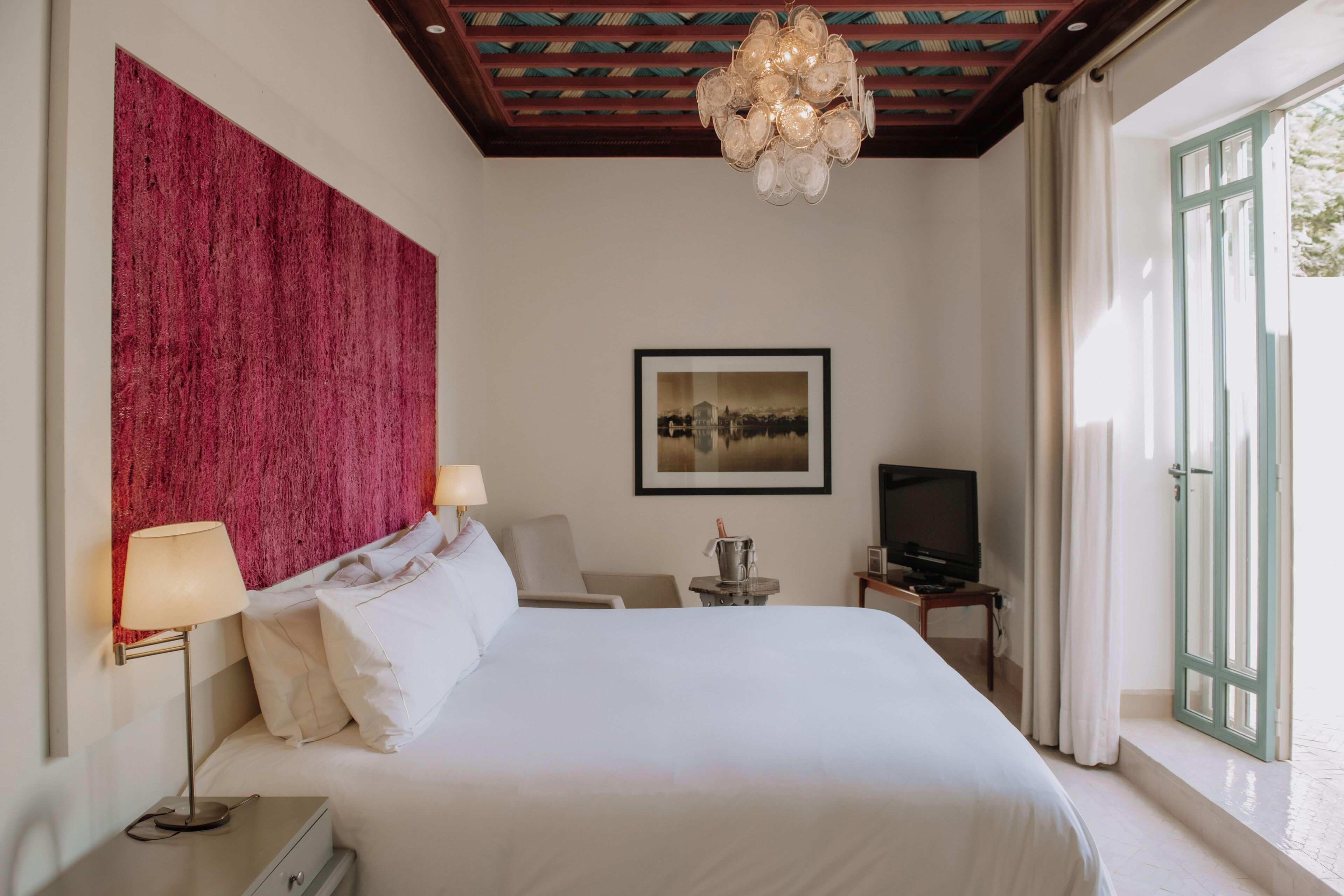 Quarto de hotel com uma cama grande, televisão, escrivaninha com luminária e vista para o exterior pela janela.