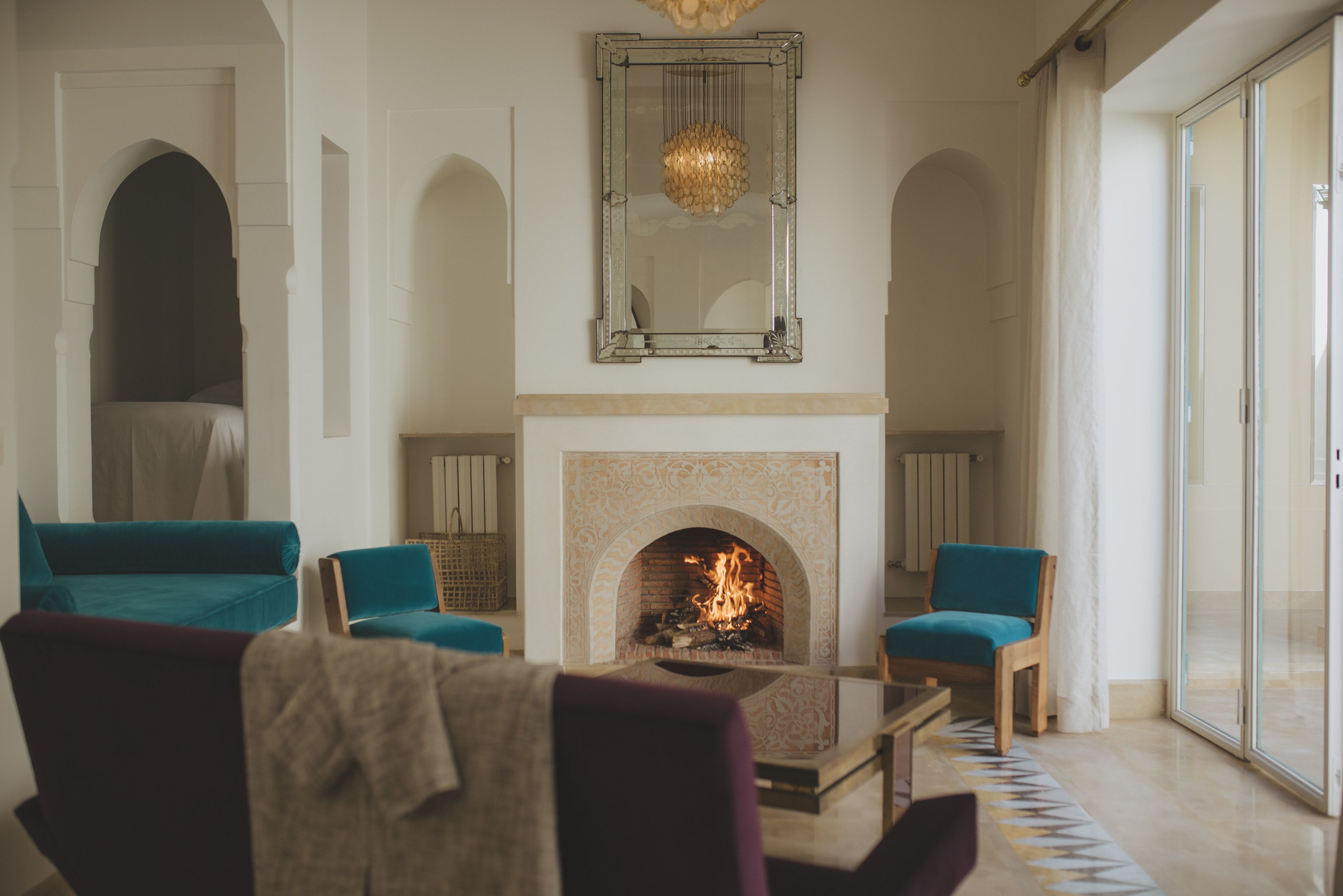 A living room with a fireplace, two teal armchairs, a coffee table, and a large mirror above the fireplace.