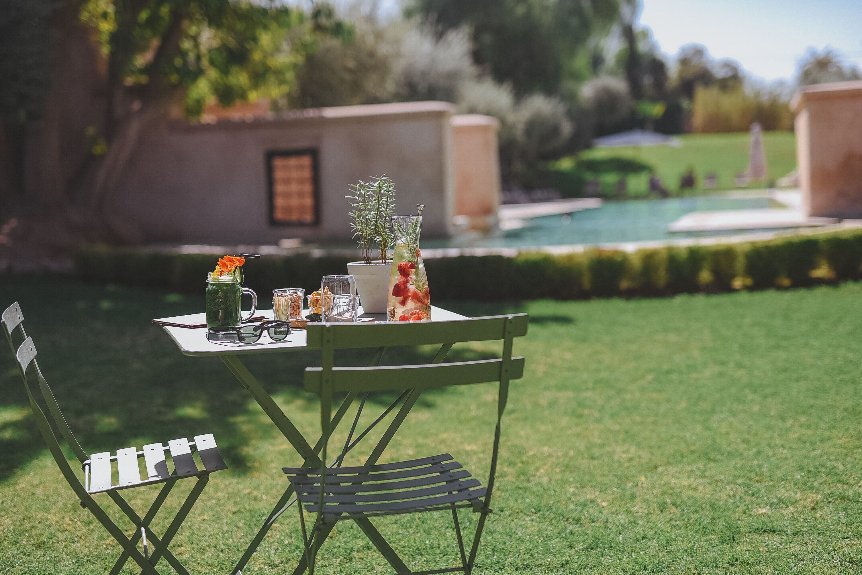 An outdoor dining area with a table set for two, surrounded by a well-manicured lawn and a pool in the background.