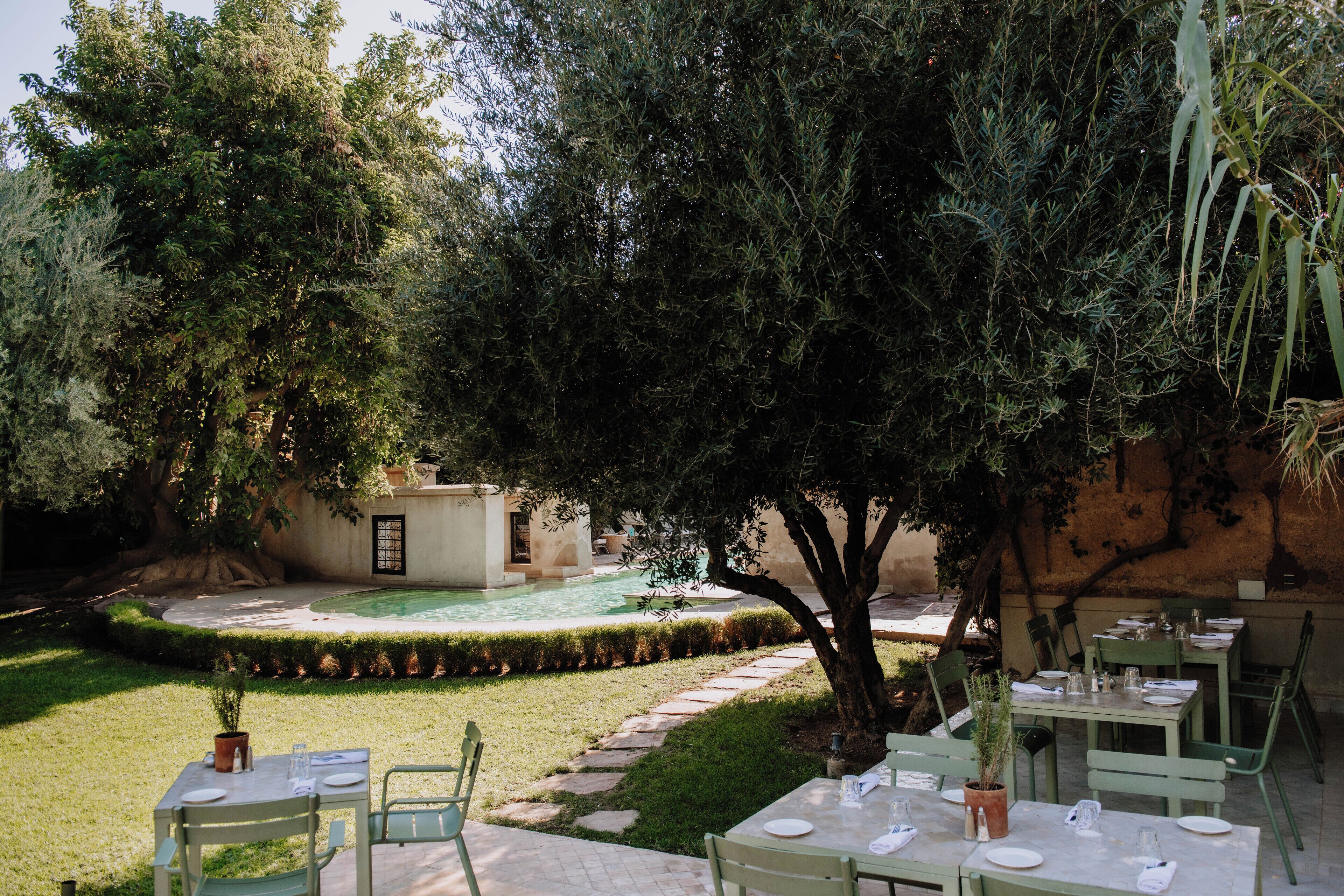 An outdoor dining area with tables and chairs, a pool, and a garden with trees and a hedge.