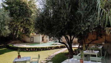 An outdoor dining area with tables and chairs, a pool, and a garden with trees and a hedge.