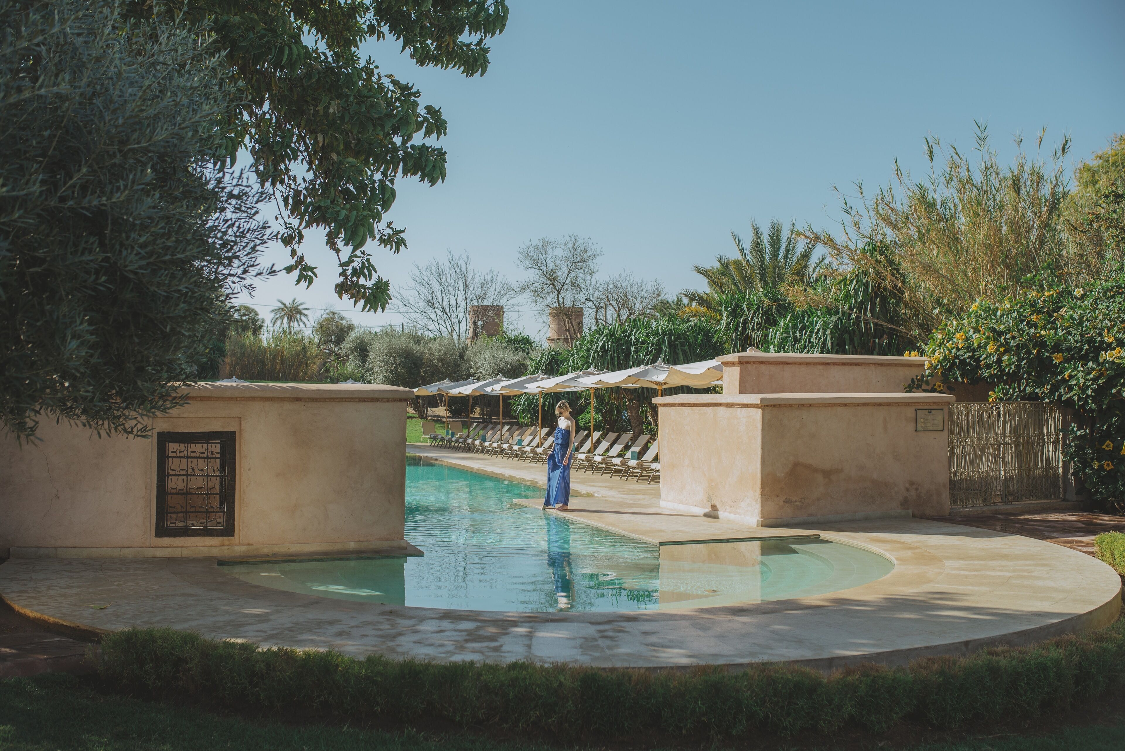 Un área de piscina con una persona vestida de azul, rodeada de follaje y un cielo despejado.