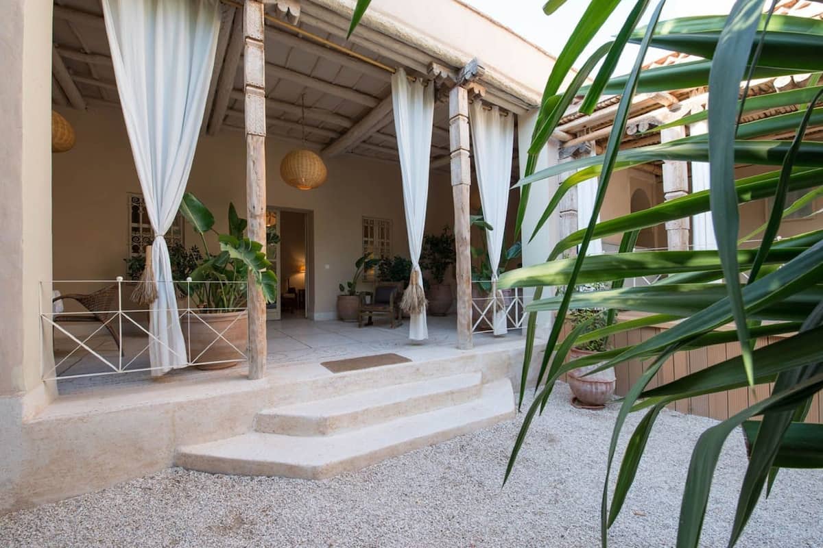 A covered outdoor area with steps leading to a building entrance, surrounded by potted plants and a white fence.