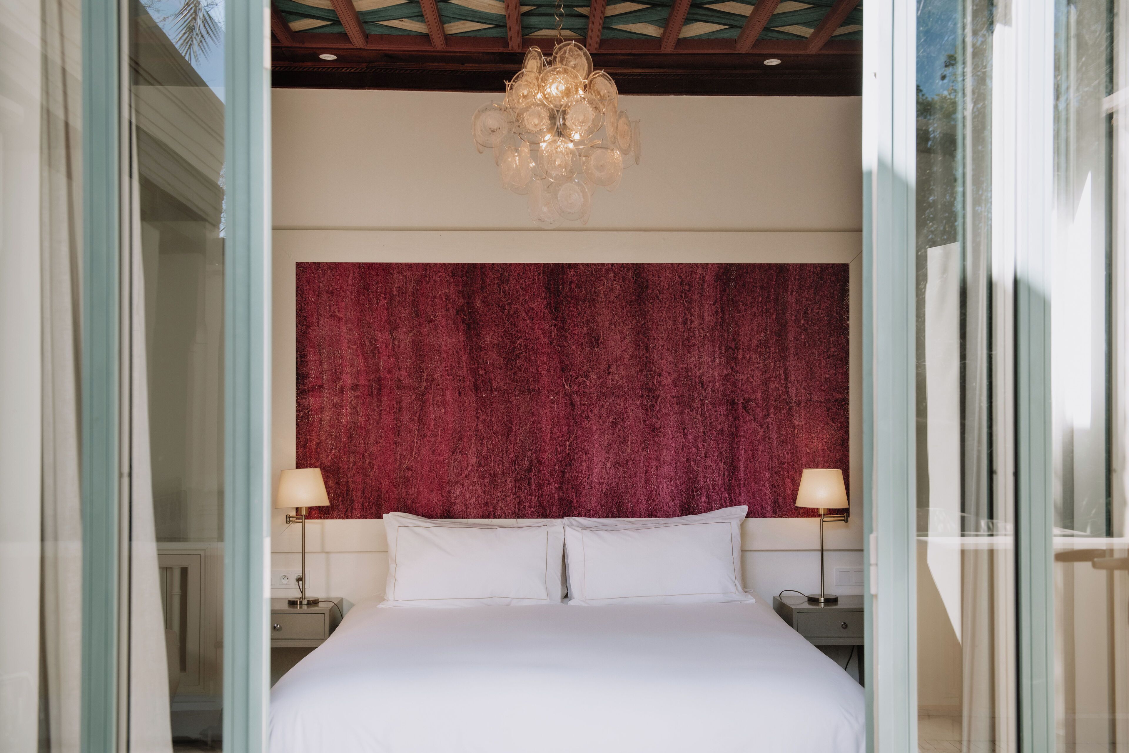 A bedroom with a large bed, two bedside tables with lamps, a red textured headboard, and a chandelier.