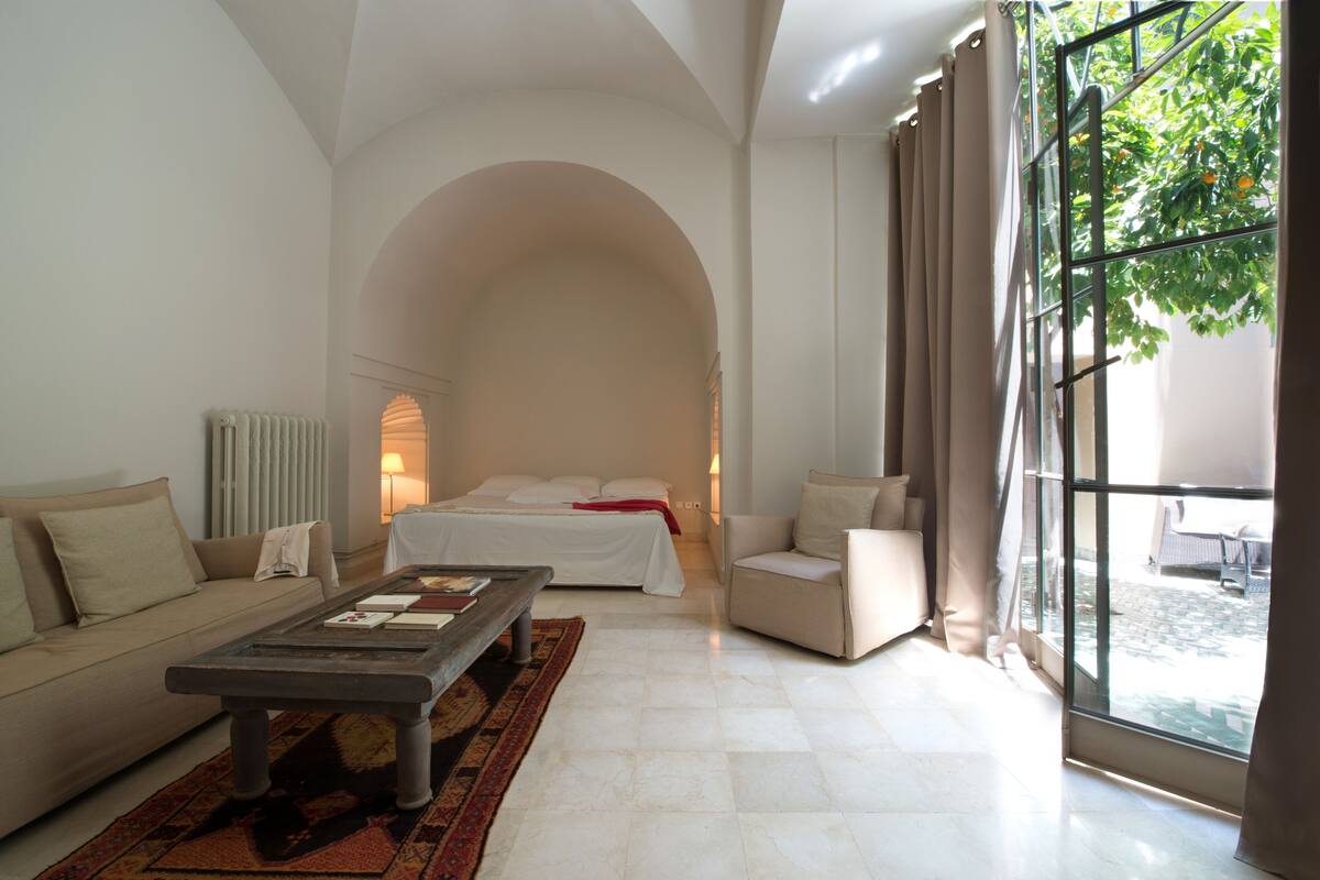 A modern hotel room with a large arched doorway, a bed with white linens, a sofa, a coffee table, and a view of the outdoors through a large window.