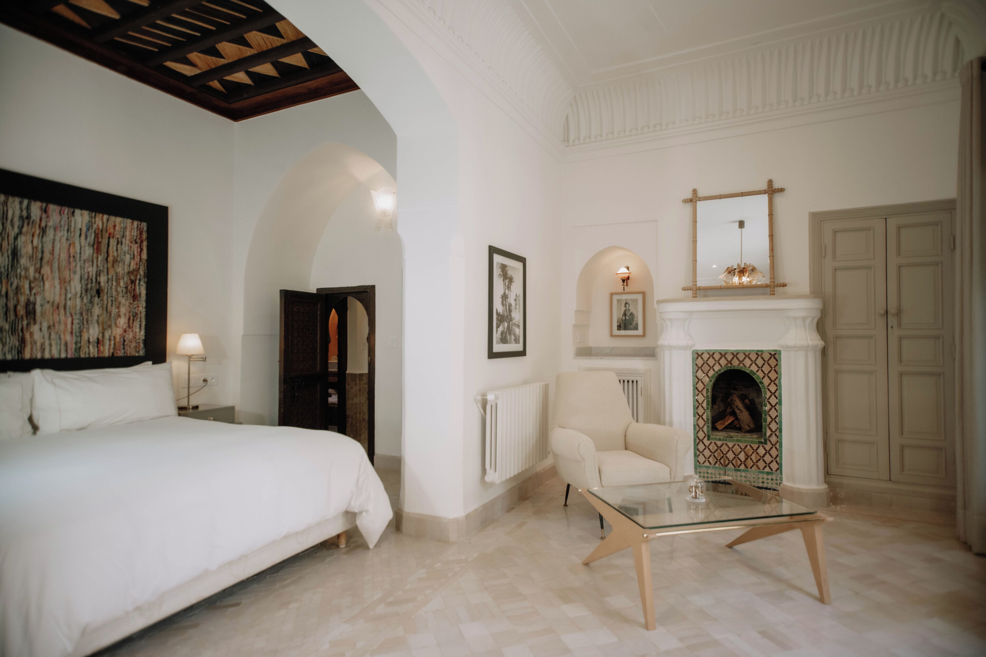 A hotel room with a bed, a chair, a glass coffee table, a fireplace, and a door.
