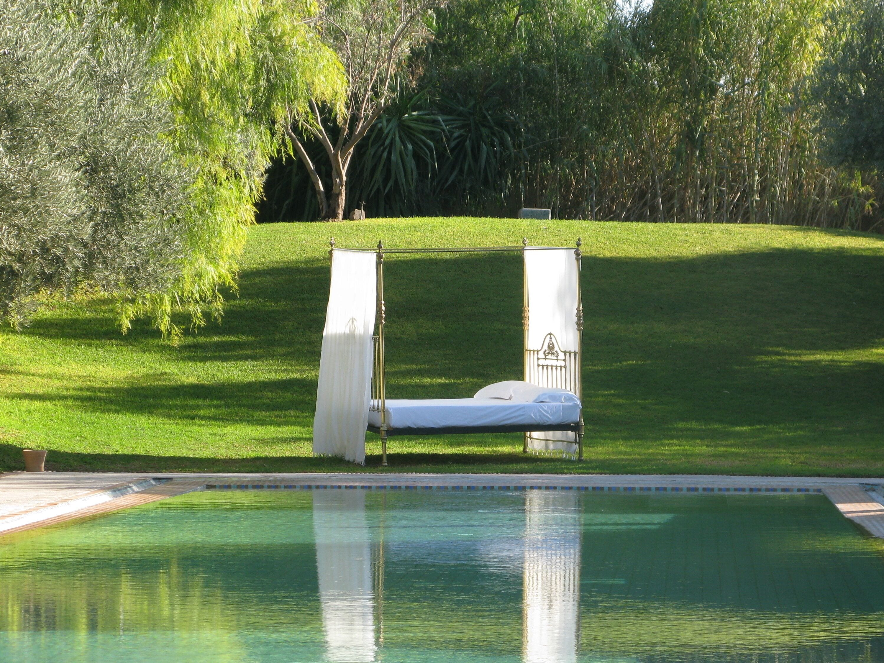 Un lit installé au bord de la piscine, surmonté d’un auvent, et entouré d’une pelouse bien entretenue et d’arbres.