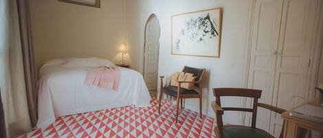 A hotel room with a bed, a chair, a desk, a mirror, and a painting on the wall.