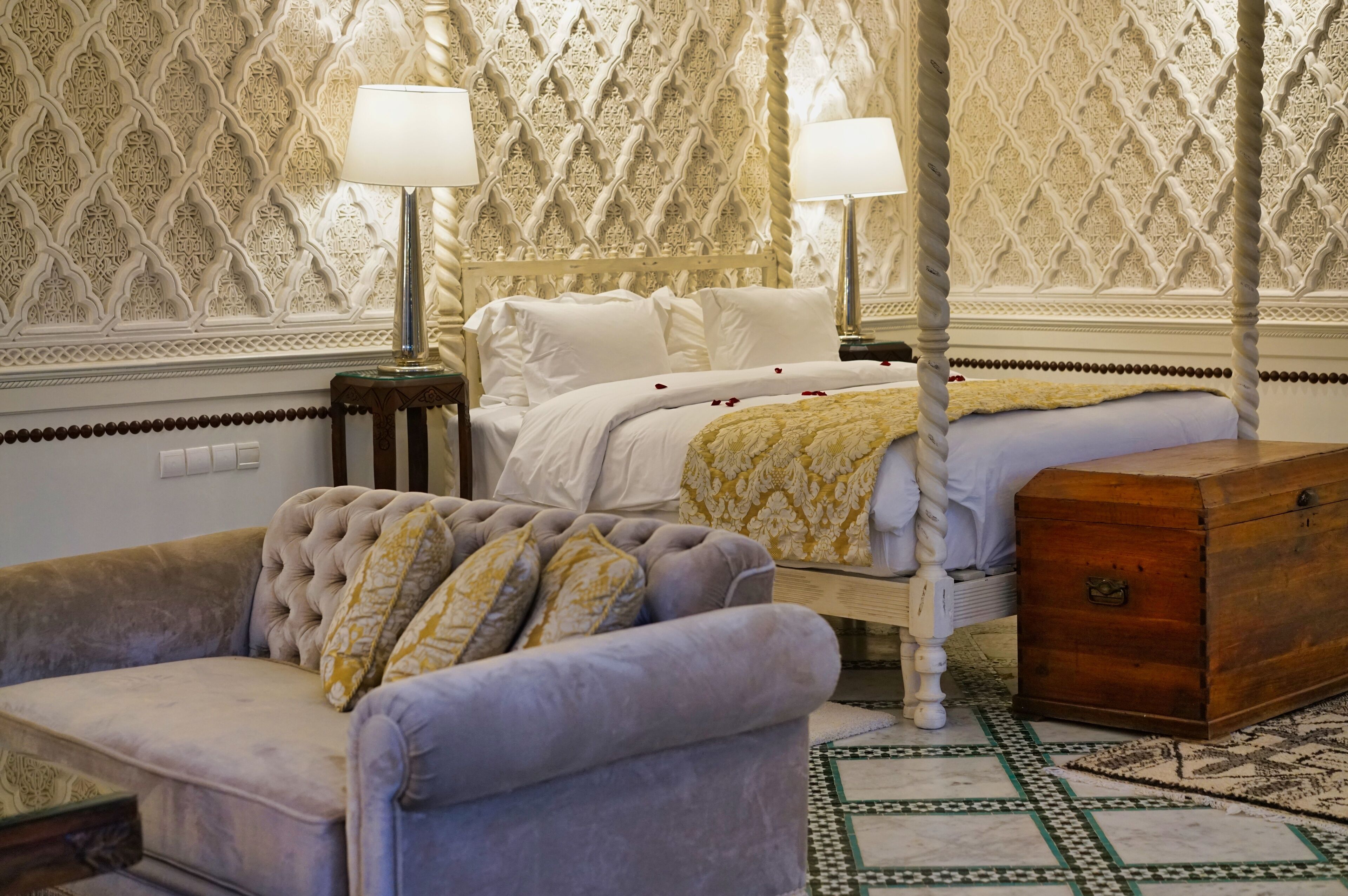 A four-poster canopy bed with white linens, a wooden chest, and a tufted sofa with patterned cushions.
