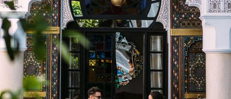 Una pareja sentada a una mesa junto a una piscina, con detalles arquitectónicos ornamentales y un ventanal grande.