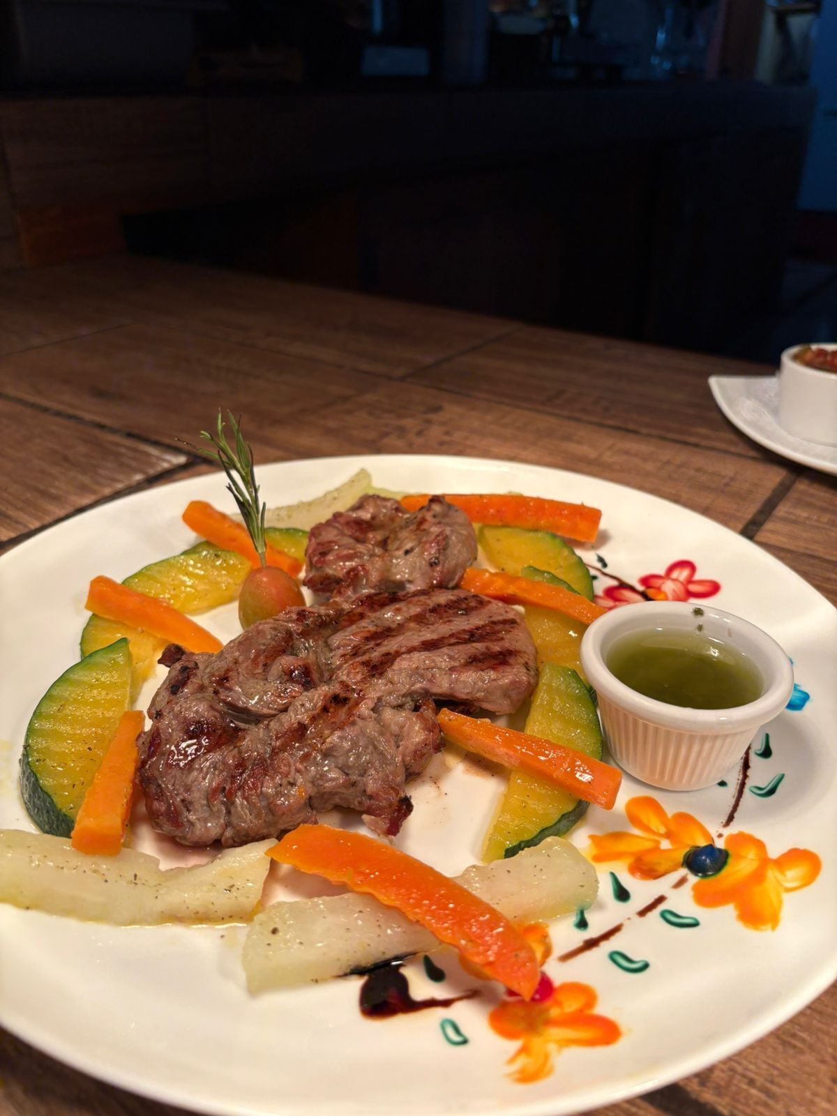 A plate of grilled meat with vegetables and a small bowl of sauce.