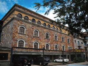 A multi-story building with a stone facade, arched windows, and a black metal fence in front. - Hotel Plaza Real (San José)
