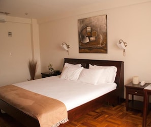 Un lit bien fait, avec du linge de lit blanc et un plaid marron, une tête de lit en bois, une table de chevet avec une lampe, et une œuvre d’art accrochée au mur au-dessus du lit.