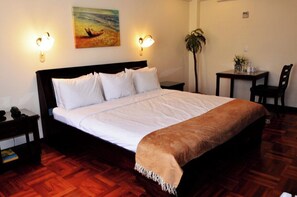 Un lit bien fait, avec du linge de lit blanc et un plaid marron, une table de chevet avec une lampe, une petite table avec une plante et un tableau accroché au mur.