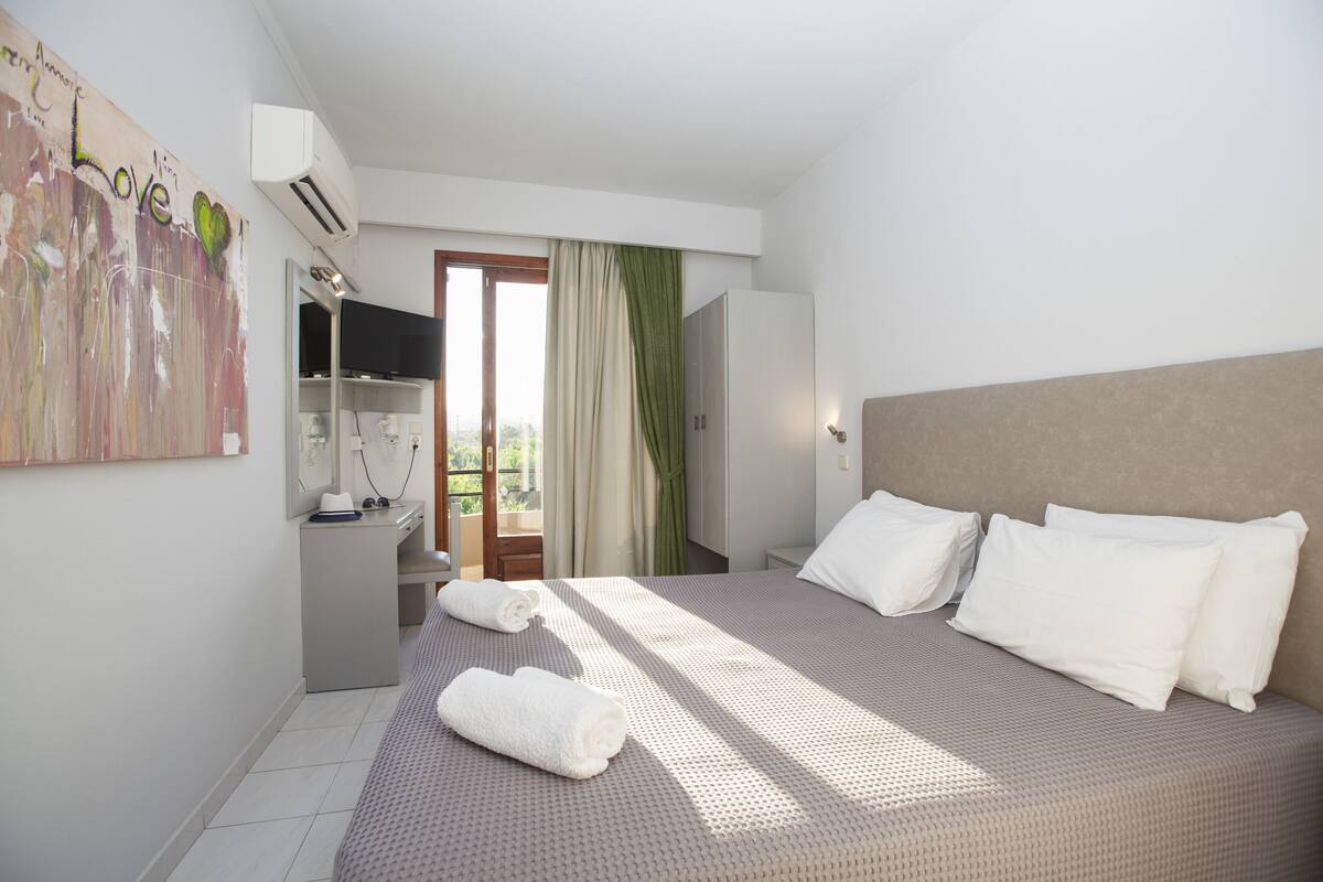 A hotel room with a bed, a desk, a TV, and a balcony with curtains.