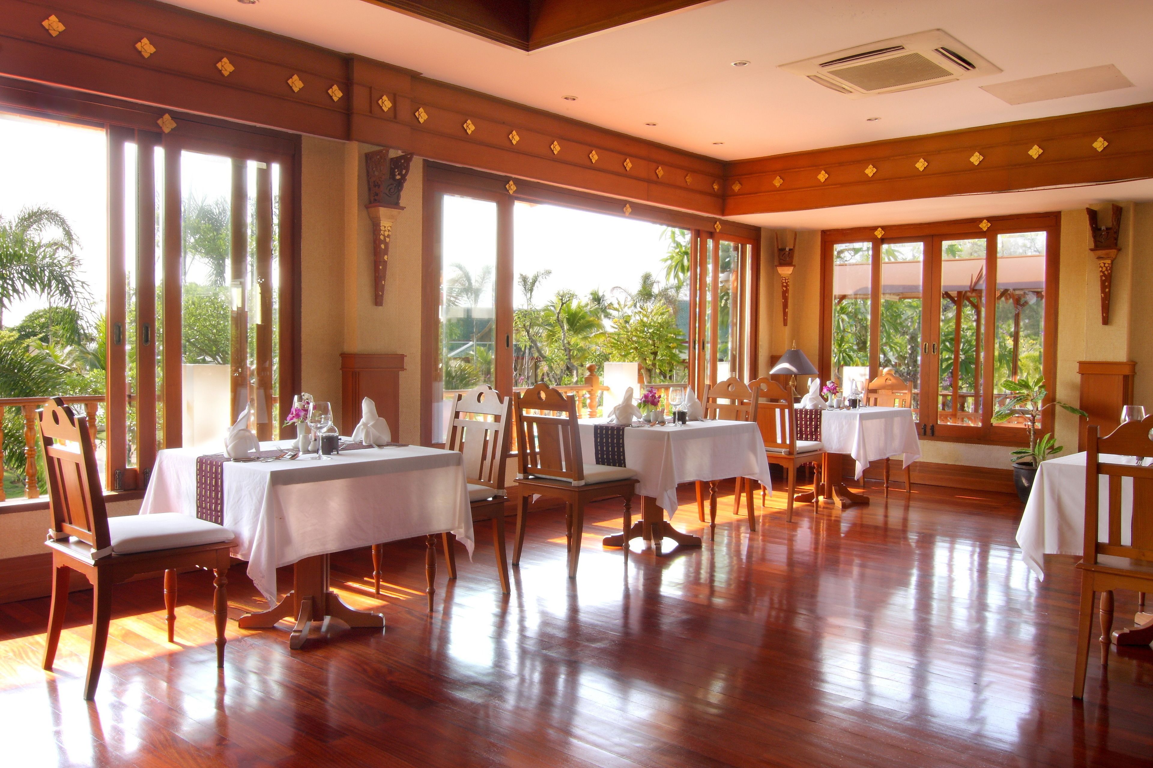 Comedor con suelo de madera, mesas y sillas blancas, y ventanas amplias con vistas a zonas verdes.
