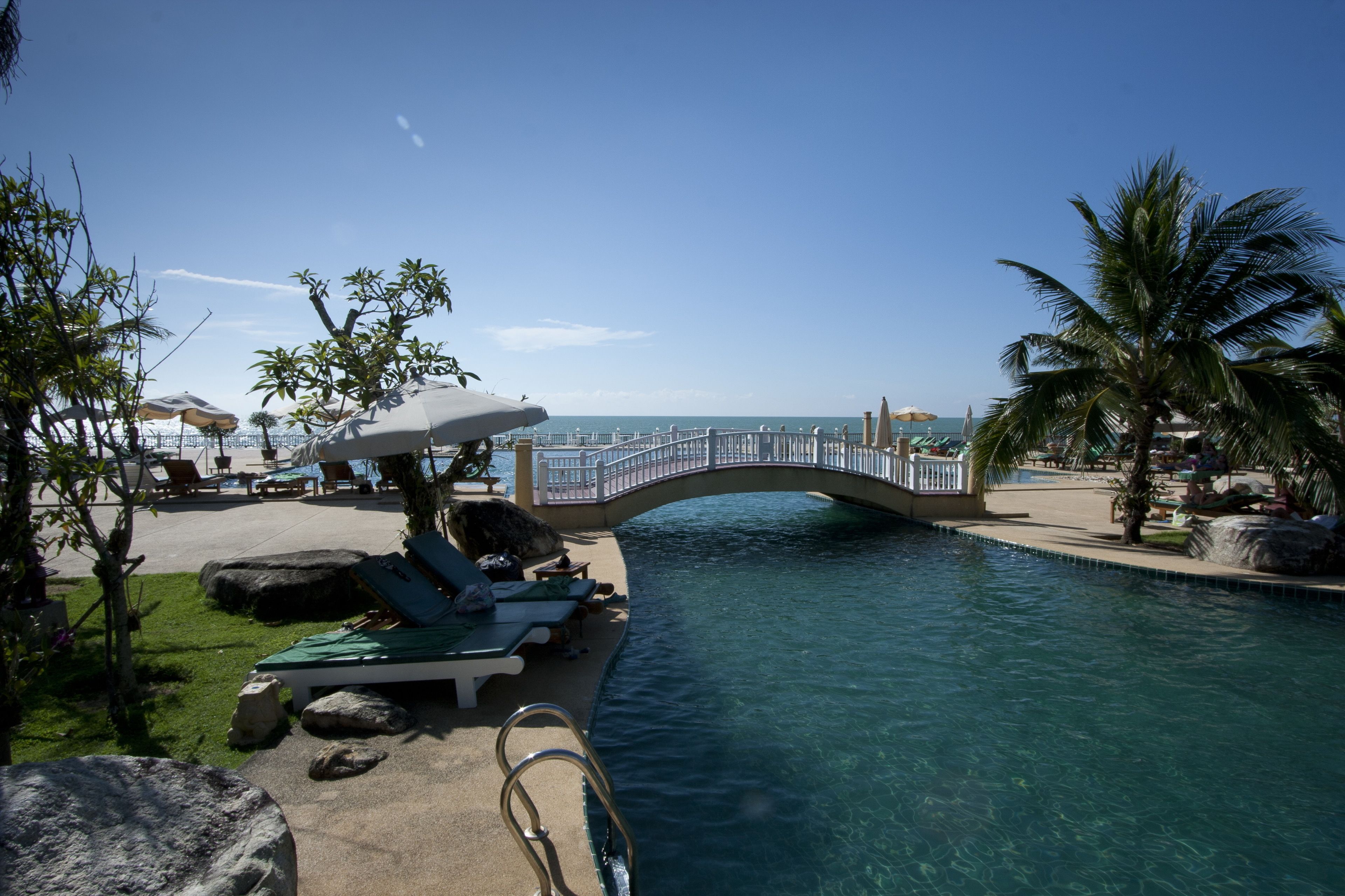 Un área de piscina con sillones de descanso y sombrillas, un puente sobre el agua y palmeras.